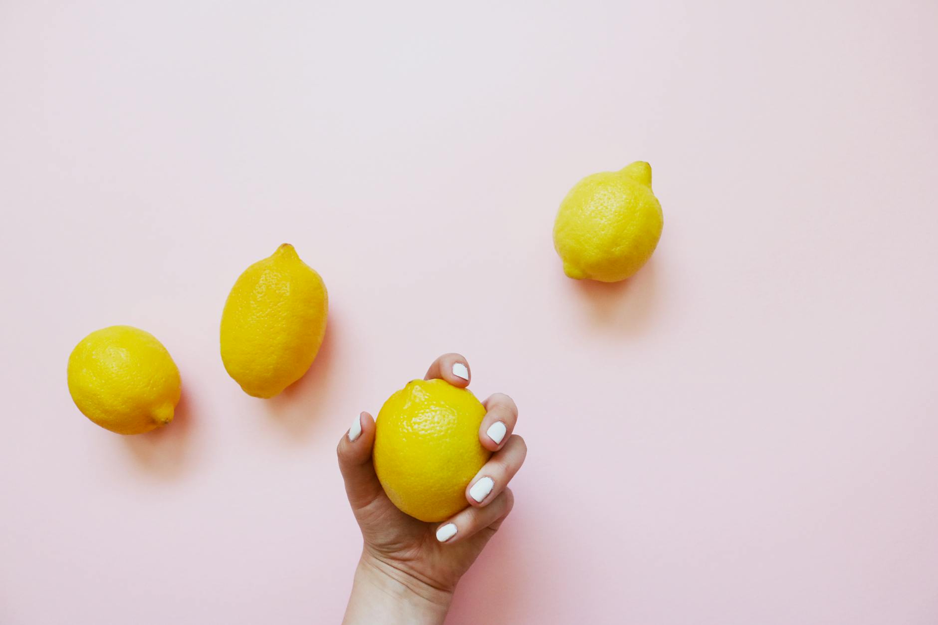 A hand holding a yellow lemon on a soft pink background surrounded by more lemons